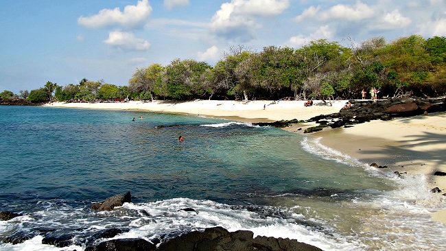 Sandy beach with turquoise water, rocky shore, trees, and scattered people under a partly cloudy sky.