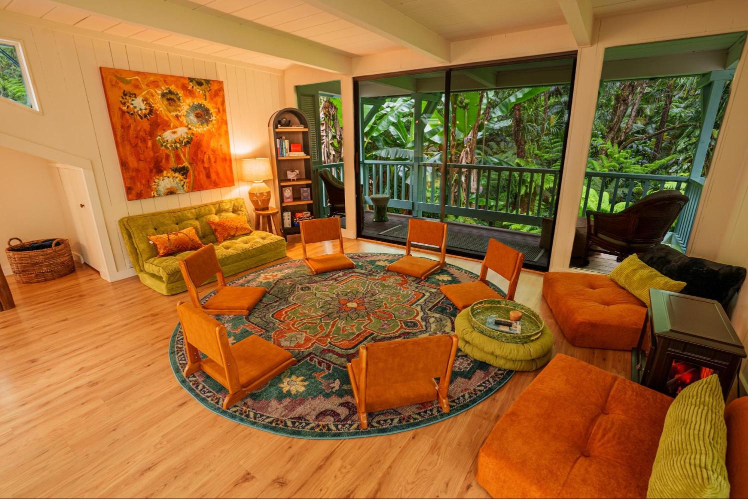 Cozy living room with orange chairs, green sofa, large rug, and lush view through windows.