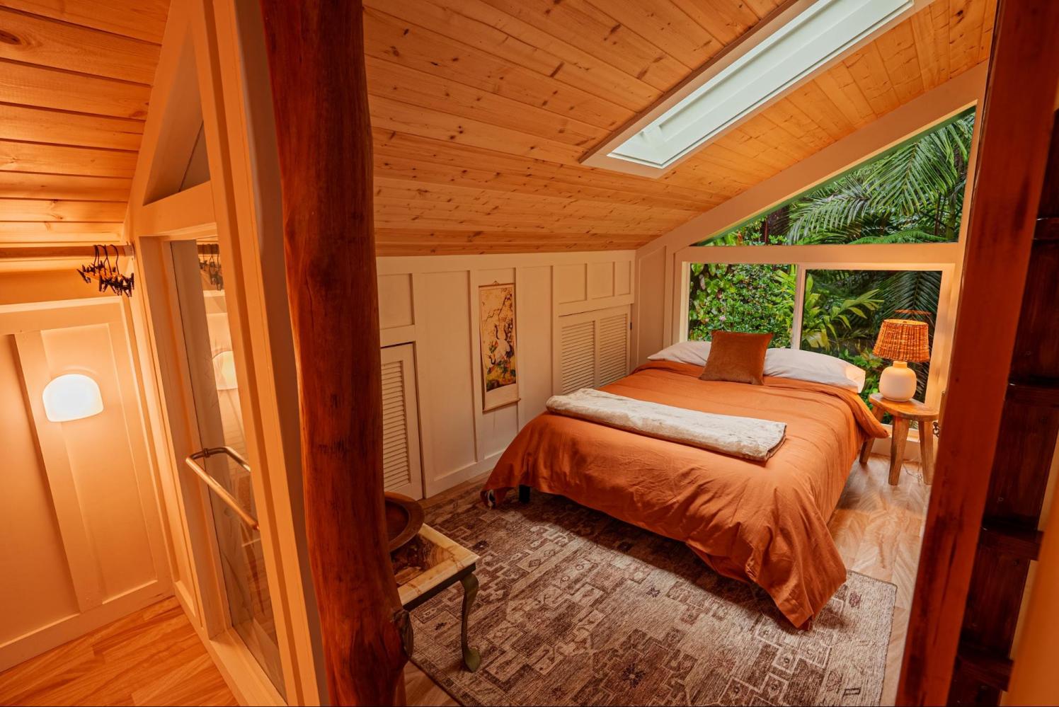 Cozy bedroom with a slanted wooden ceiling, skylight, and large window facing lush greenery.