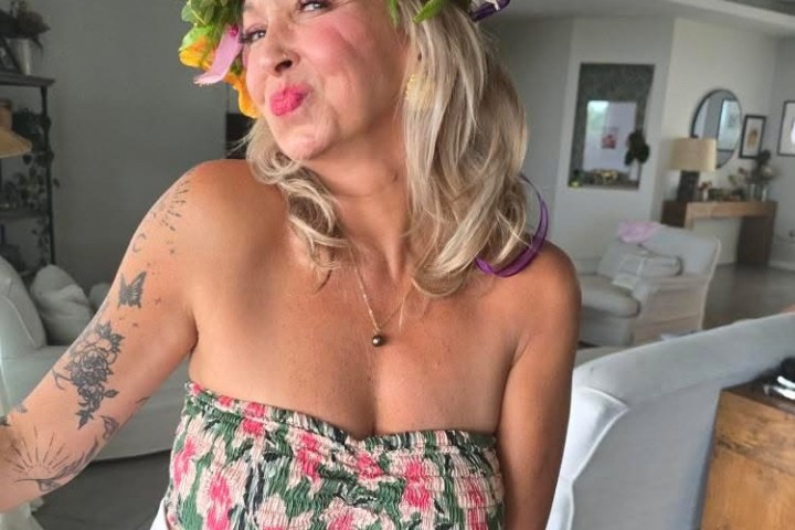 Woman indoors wearing floral top and flower crown, looking at camera with playful expression.