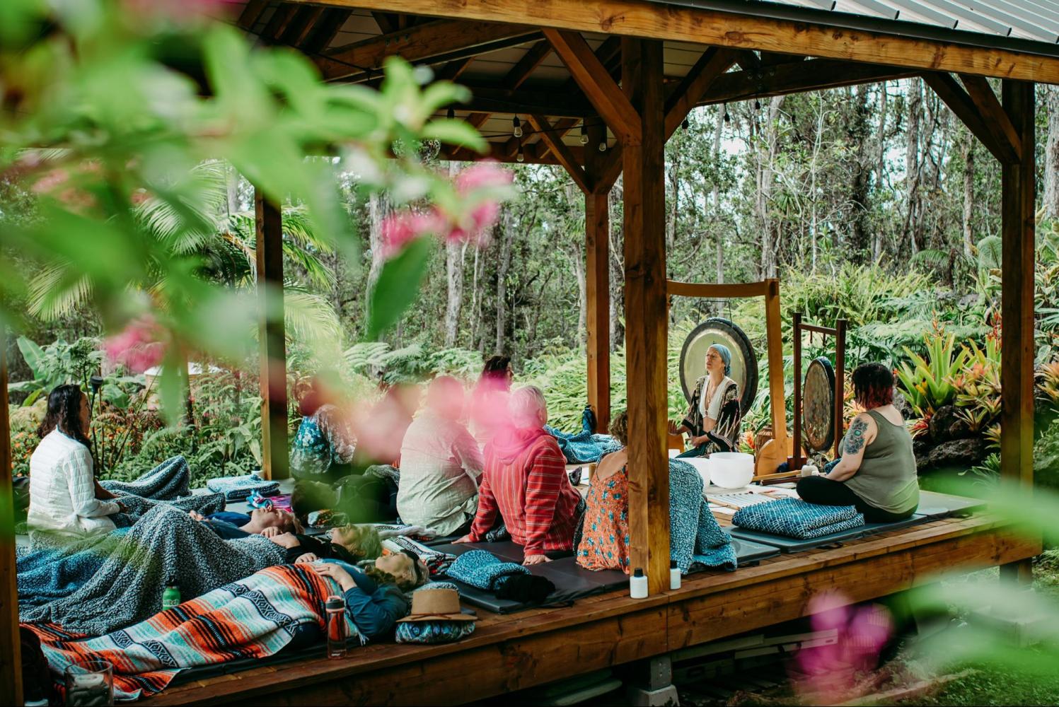 People in a wooden gazebo meditating with a gong amidst lush greenery.