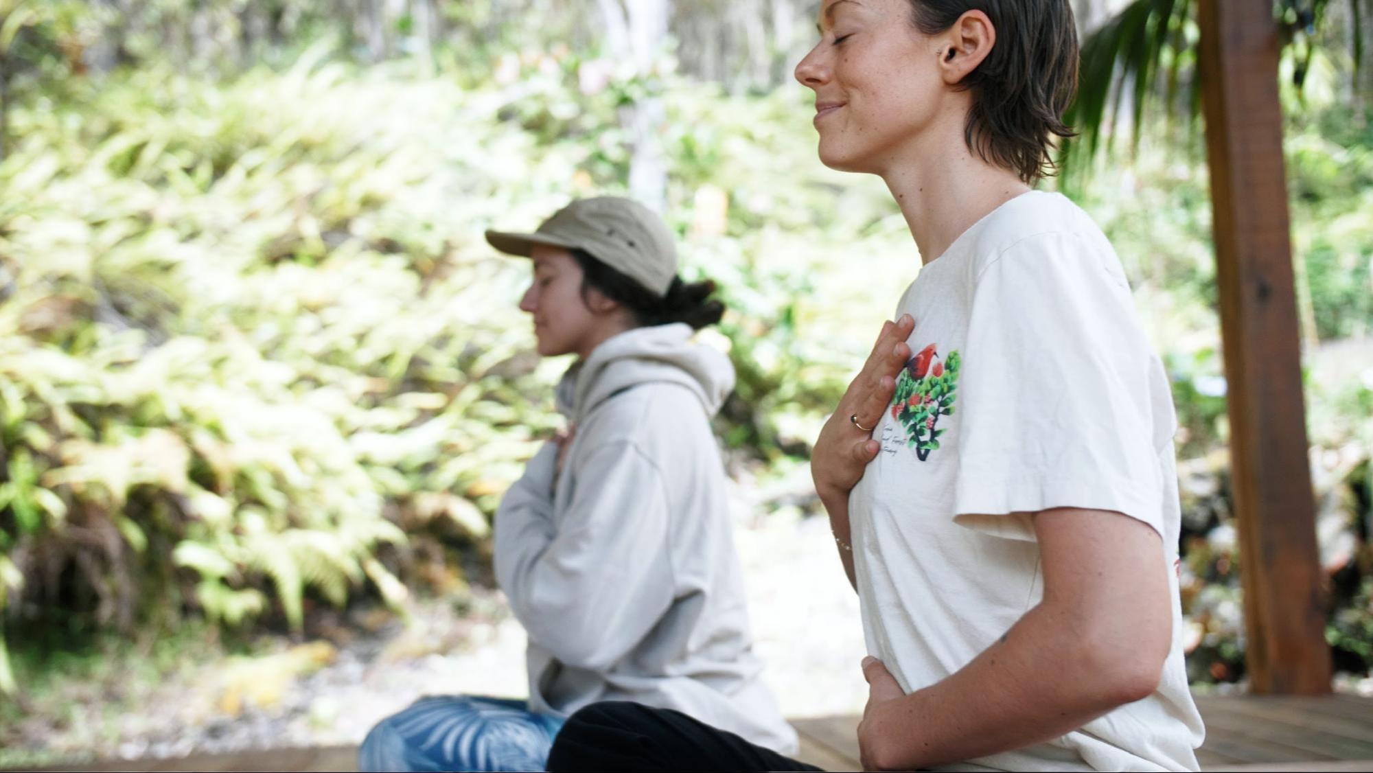Two people practicing meditation outdoors, hands on chest, with greenery in the background.