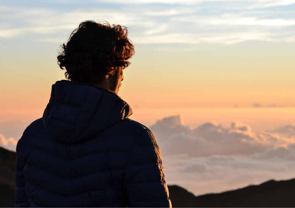 Person in a jacket gazing at a vibrant sunset over clouds from a mountain viewpoint.