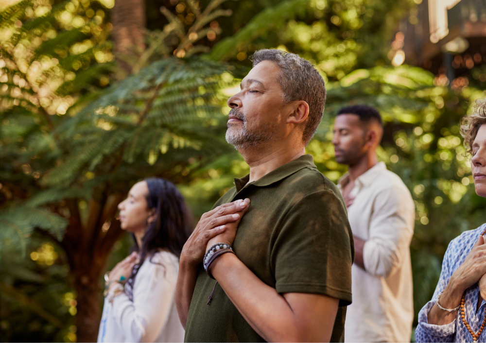 Four people meditating outdoors with hands on chests, surrounded by greenery.