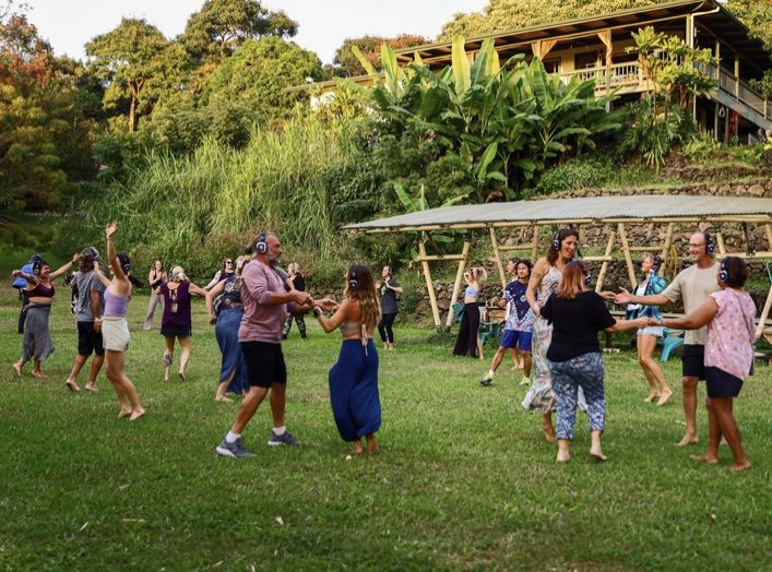 People dancing outdoors with headphones, surrounded by lush greenery and a wooden building.