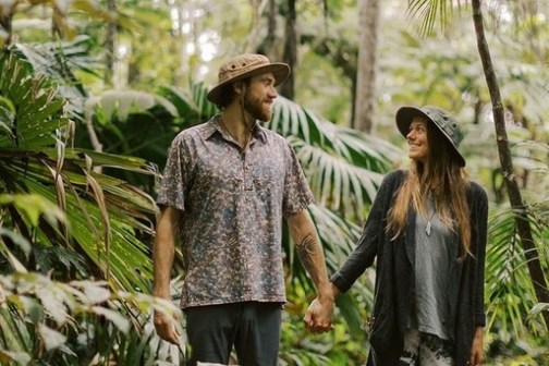 Couple holding hands in lush forest, wearing casual outdoor clothing and hats.