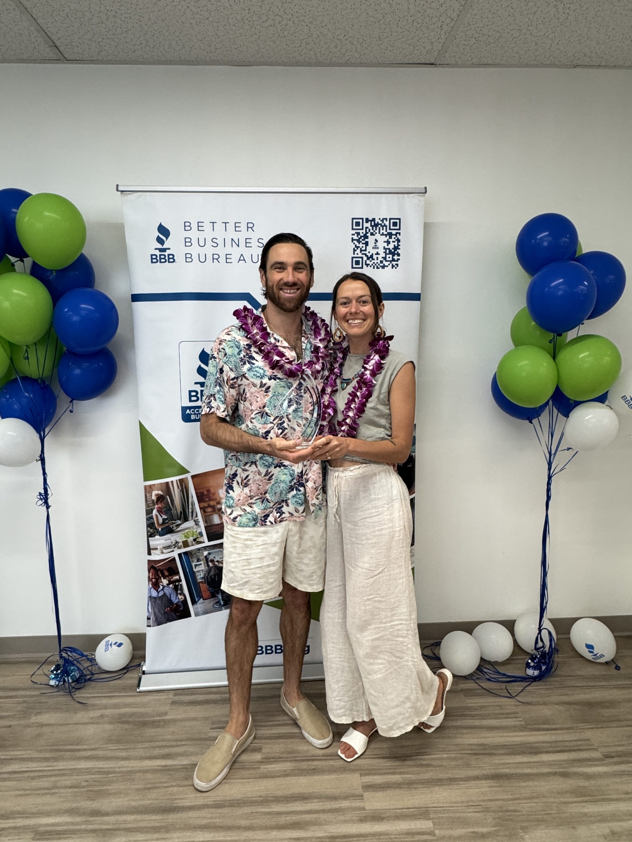 Man and woman in leis stand smiling with a Better Business Bureau backdrop and balloons.
