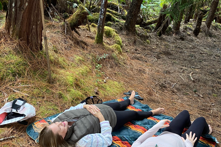 Two people relaxing on a blanket in a forest, surrounded by trees and foliage.