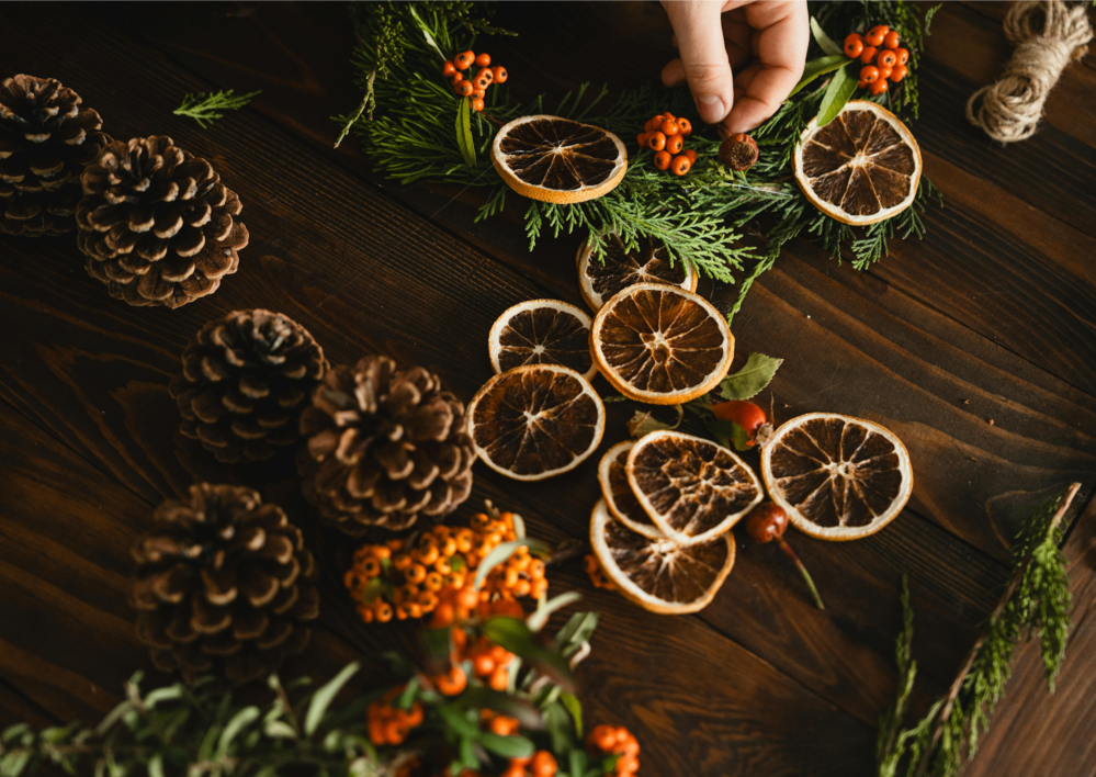 Hand arranging a wreath with dried oranges, pinecones, and berries on a wooden surface.