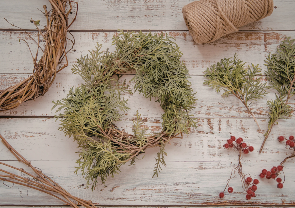 Rustic wreath making materials on a wooden surface with greenery, berries, twine, and vine wreath.