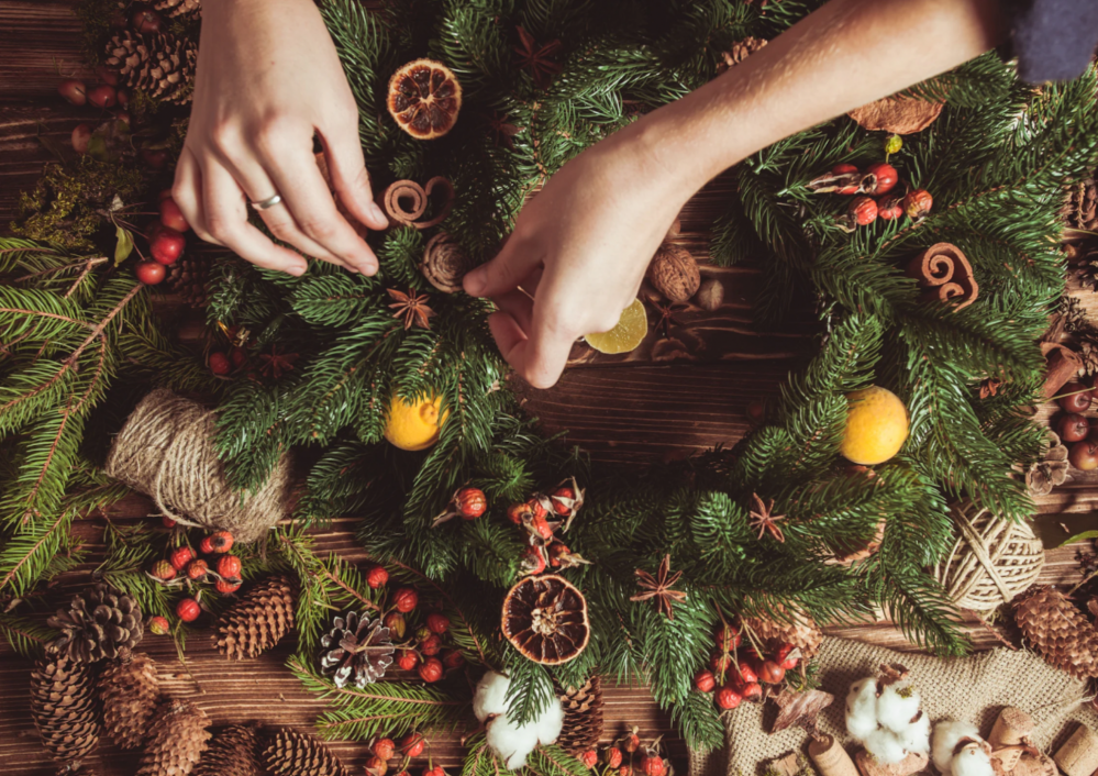 Hands decorating a wreath with pine branches, dried fruits, and cinnamon on a wooden table.