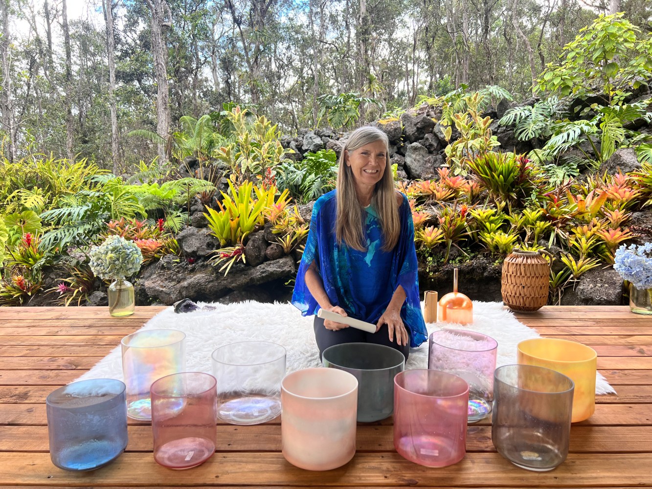 Person sitting outdoors with colorful sound bowls on a wooden deck surrounded by lush greenery.
