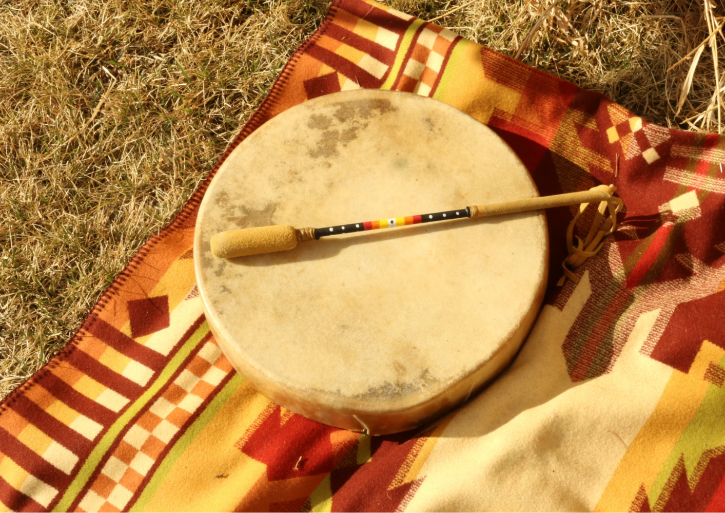 Drum and stick on a colorful patterned blanket on dry grass.