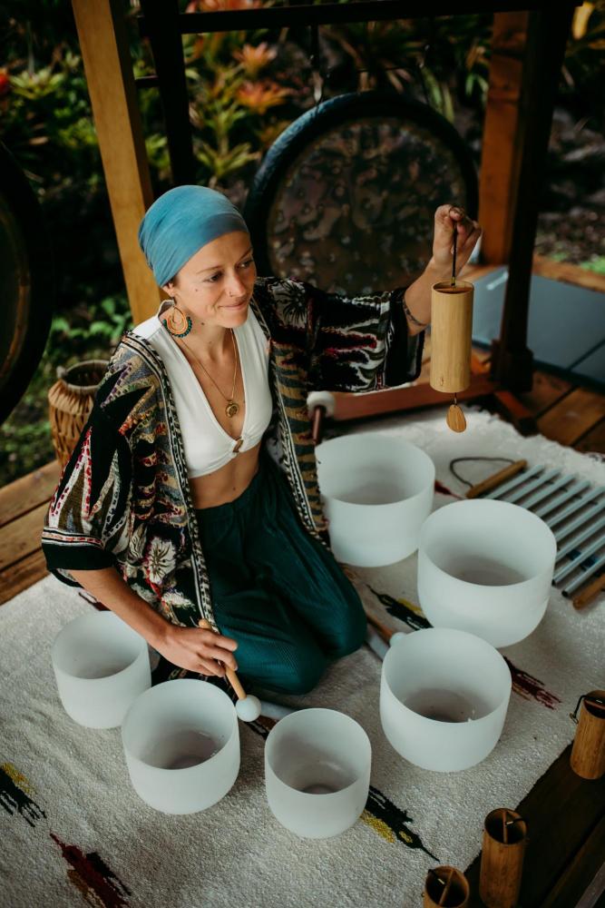 Woman playing crystal singing bowls on a rug, surrounded by serene garden setting.