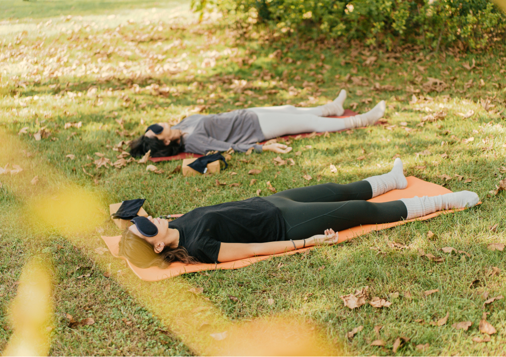 Two people lying on yoga mats outdoors wearing eye masks on a grassy area.