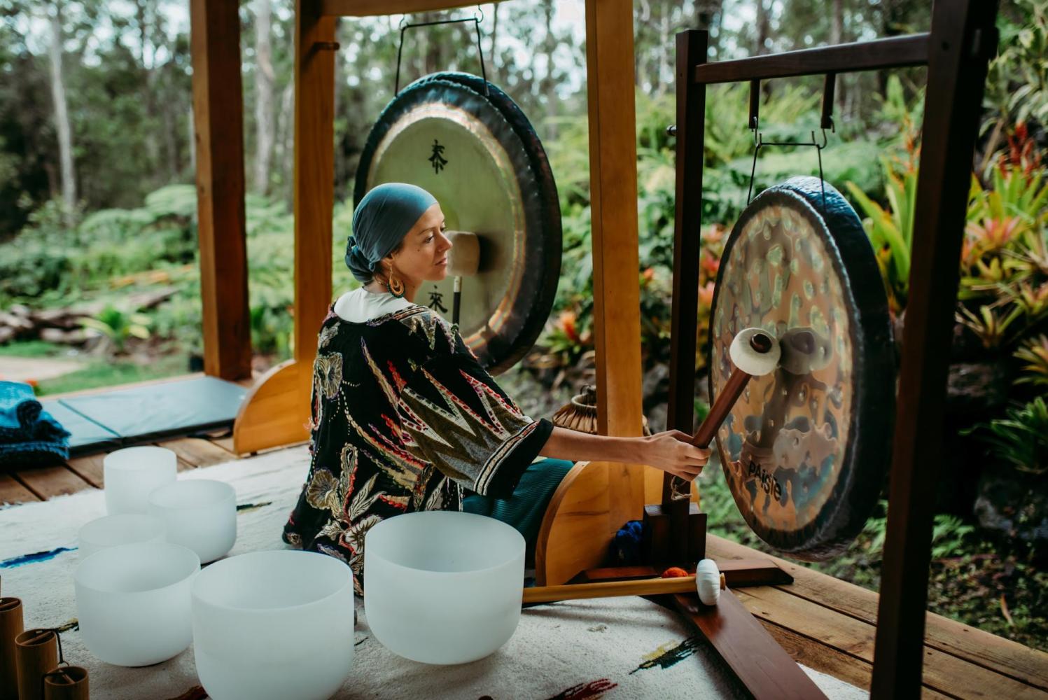 Person playing gongs, surrounded by crystal bowls on a deck with lush greenery.