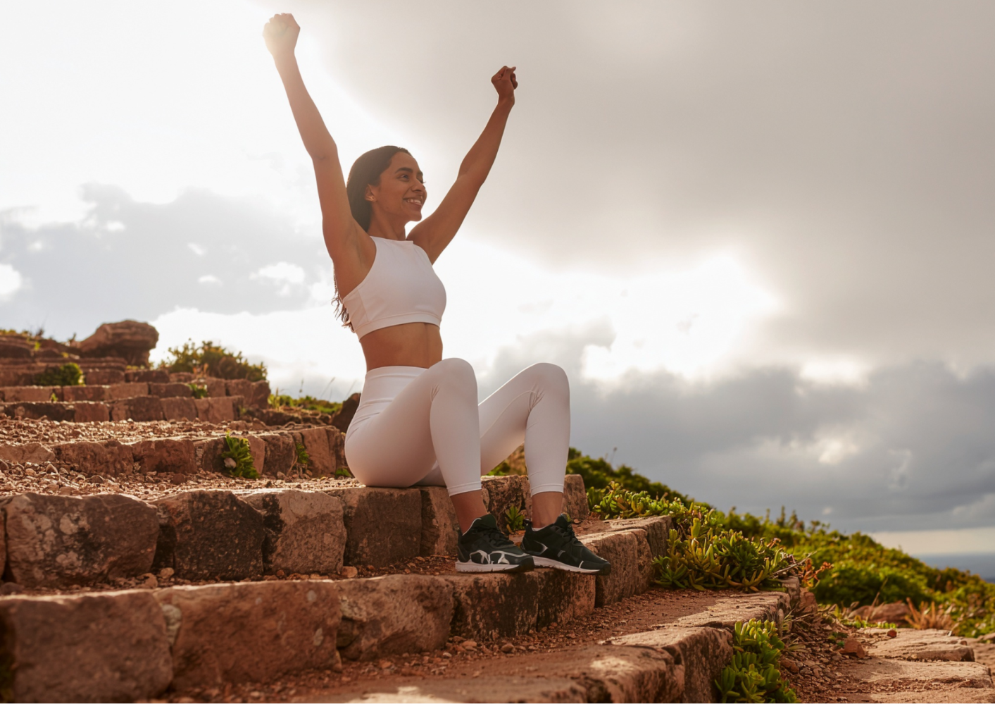 Woman in white activewear sitting on steps, arms raised, smiling, with a cloudy sky background.