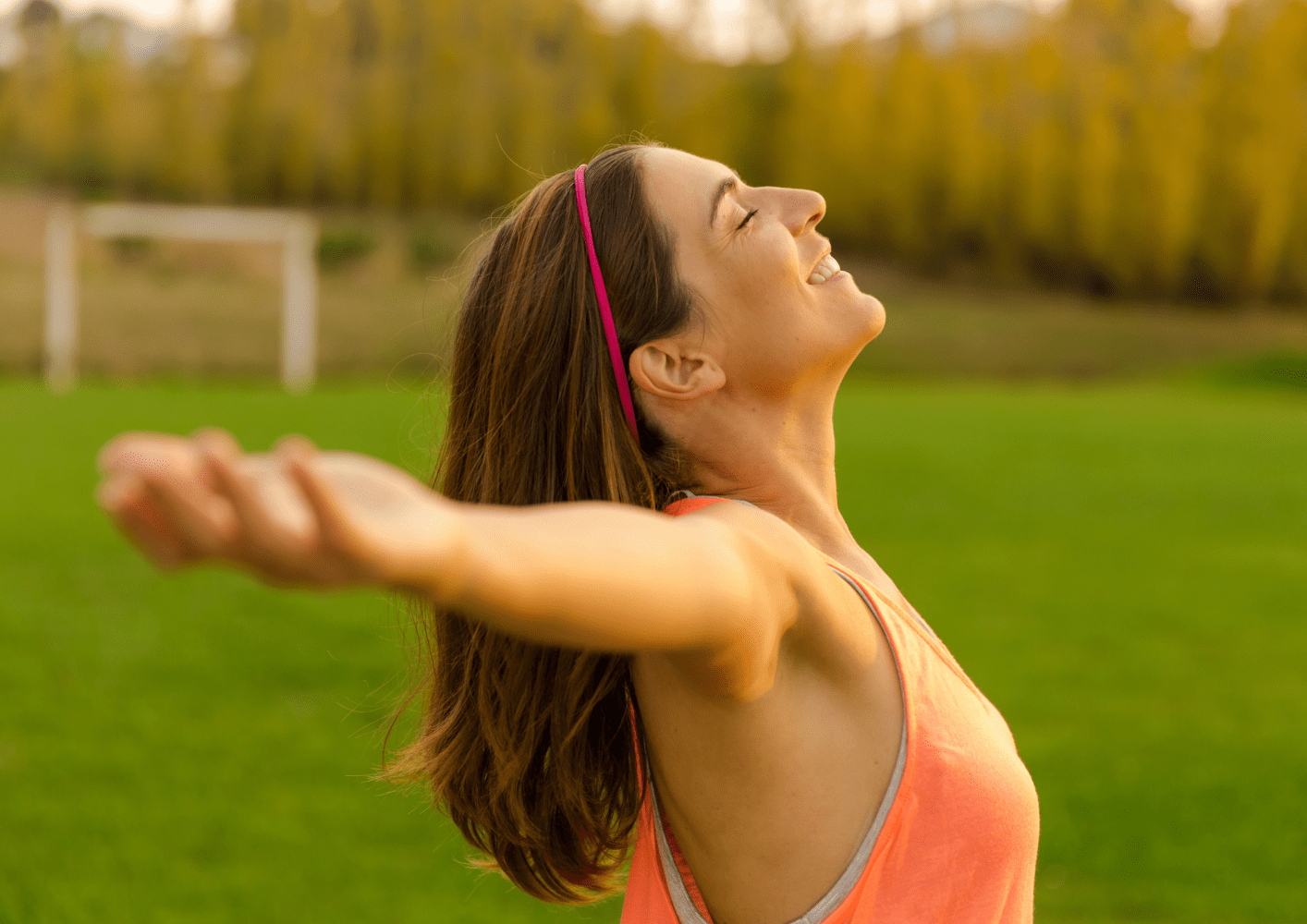 Woman in orange tank top with arms outstretched, smiling in a grassy field.