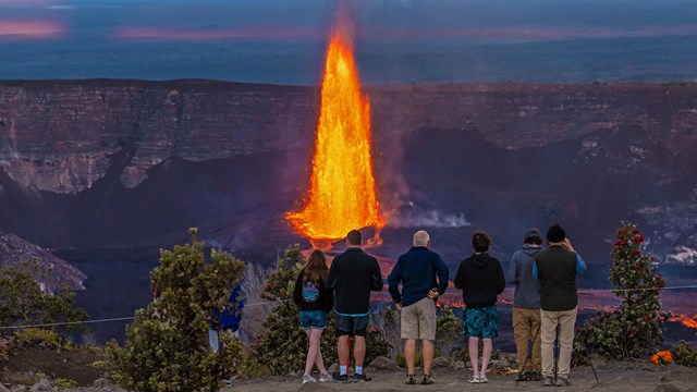 Five people watching a volcano erupt with bright orange lava at dusk.