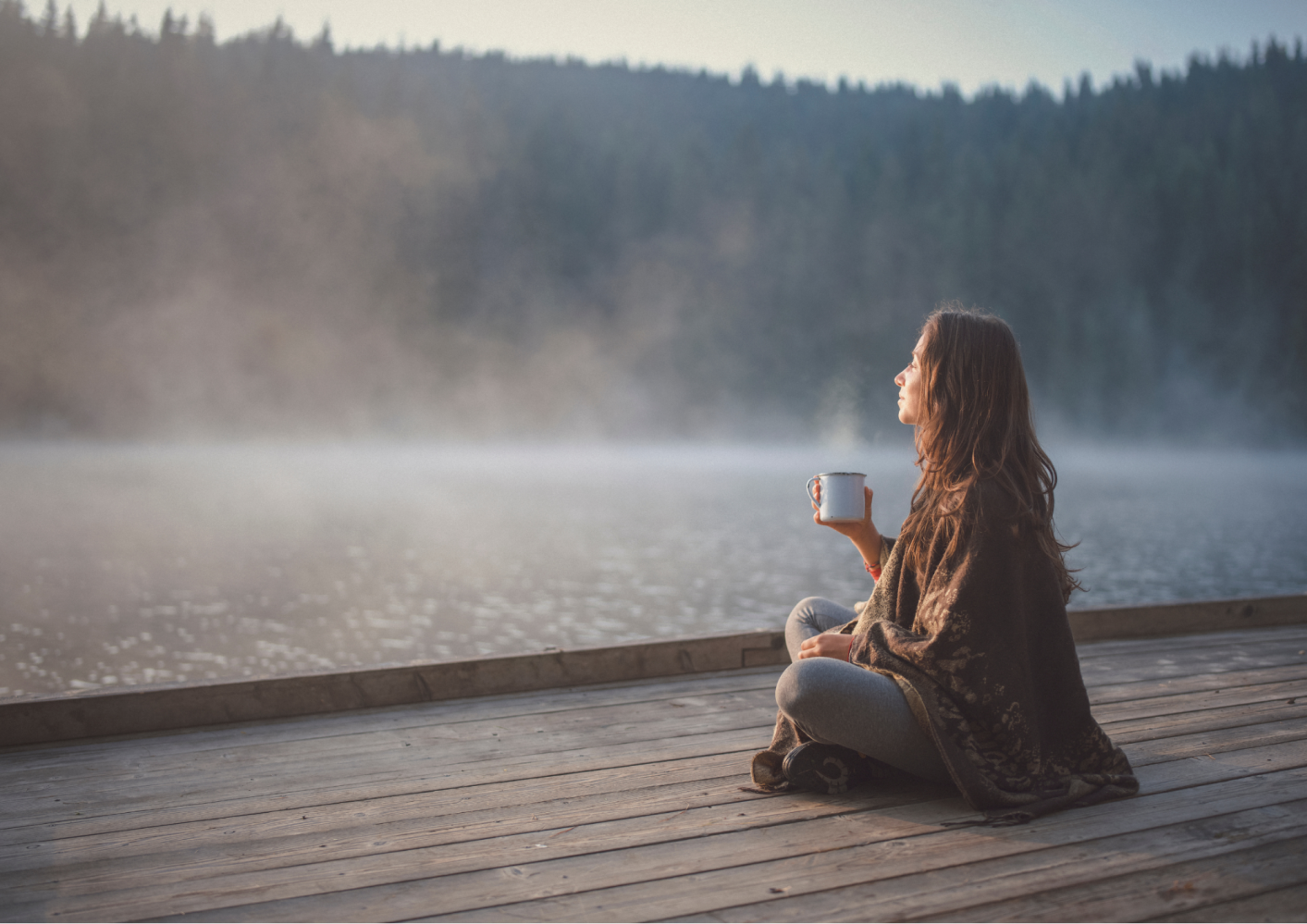 Person sitting on a dock, holding a mug, gazing at misty lake with forest background.