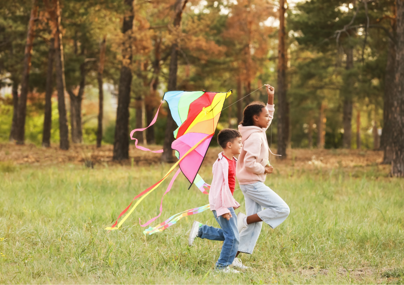 Two kids running with a colorful kite in a grassy area surrounded by trees.