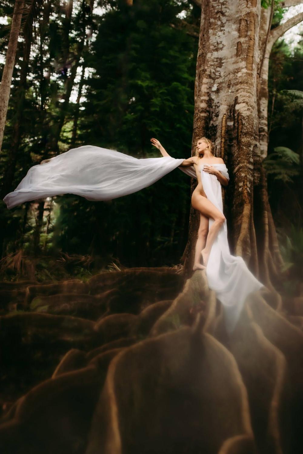 Person draped in white fabric standing on tree roots in forest.