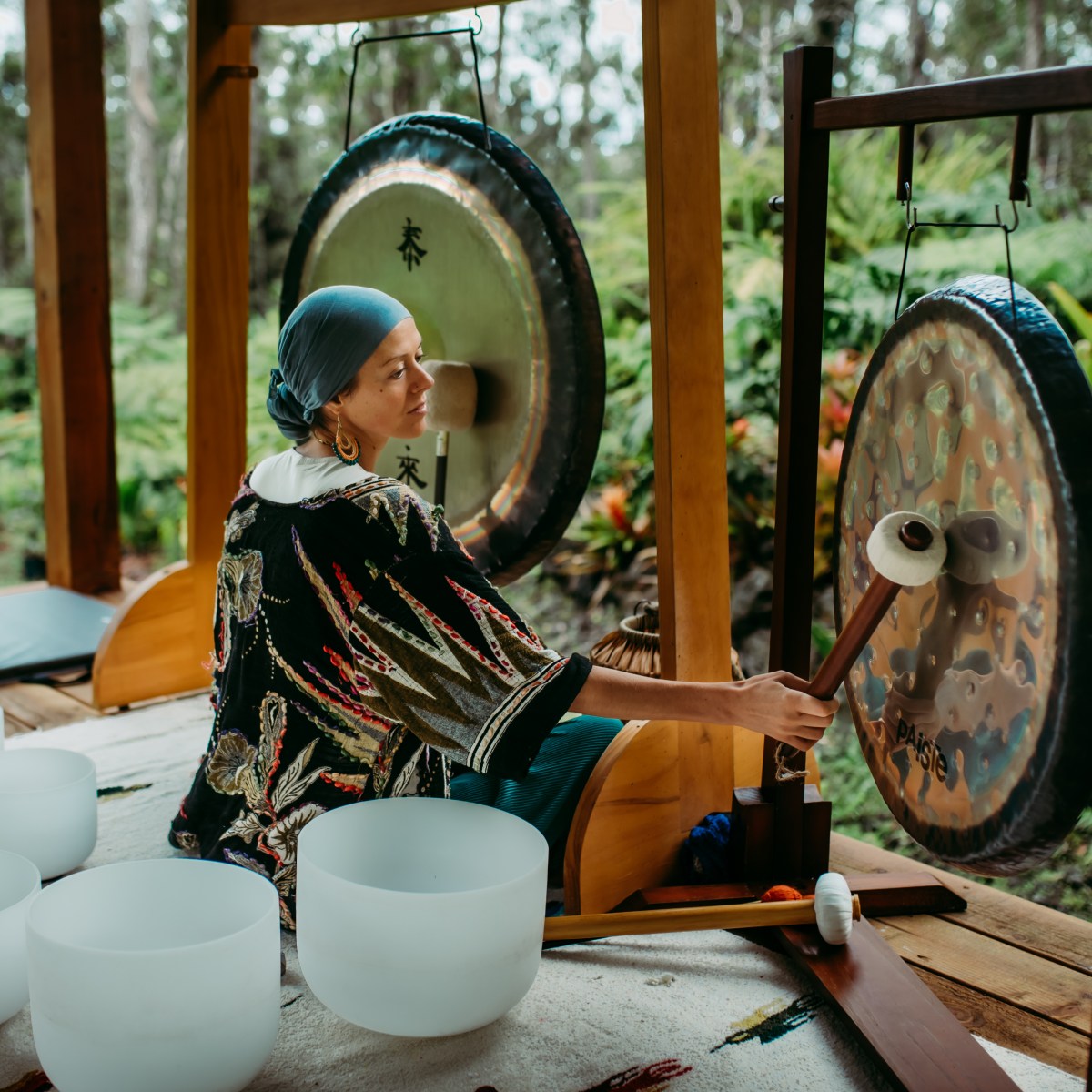 Person playing gongs in a tropical outdoor setting with singing bowls on a rug.