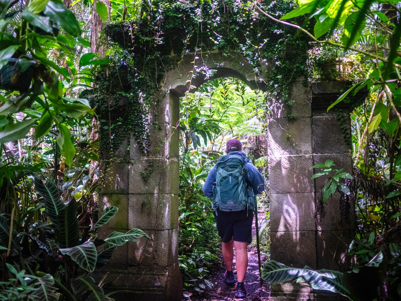Person with backpack walks through overgrown stone arch in lush jungle.
