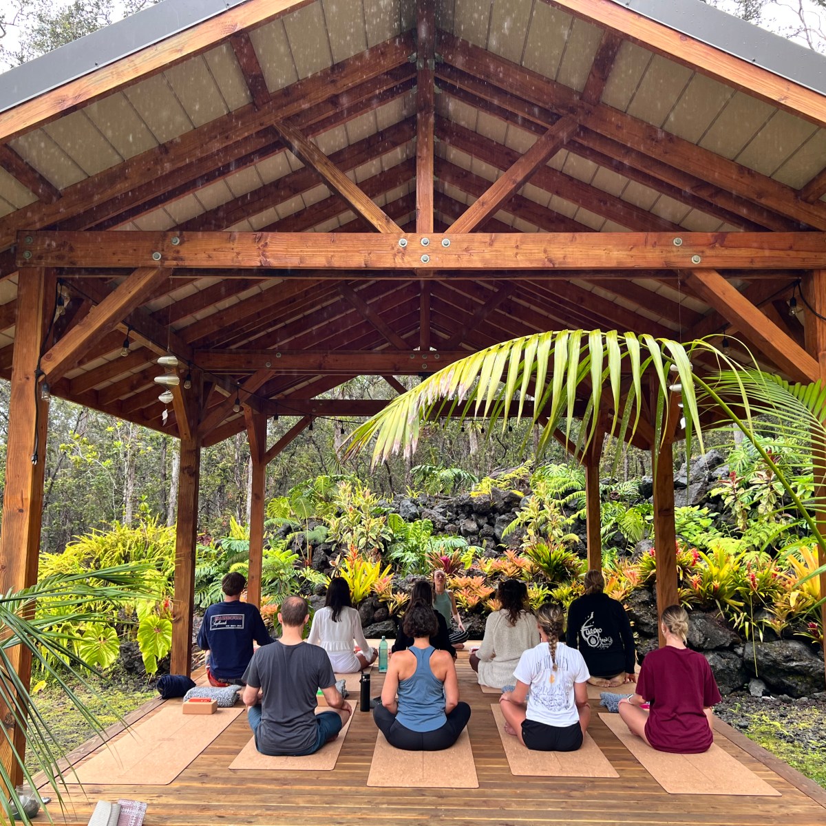 Group meditating under a wooden pavilion surrounded by lush greenery.