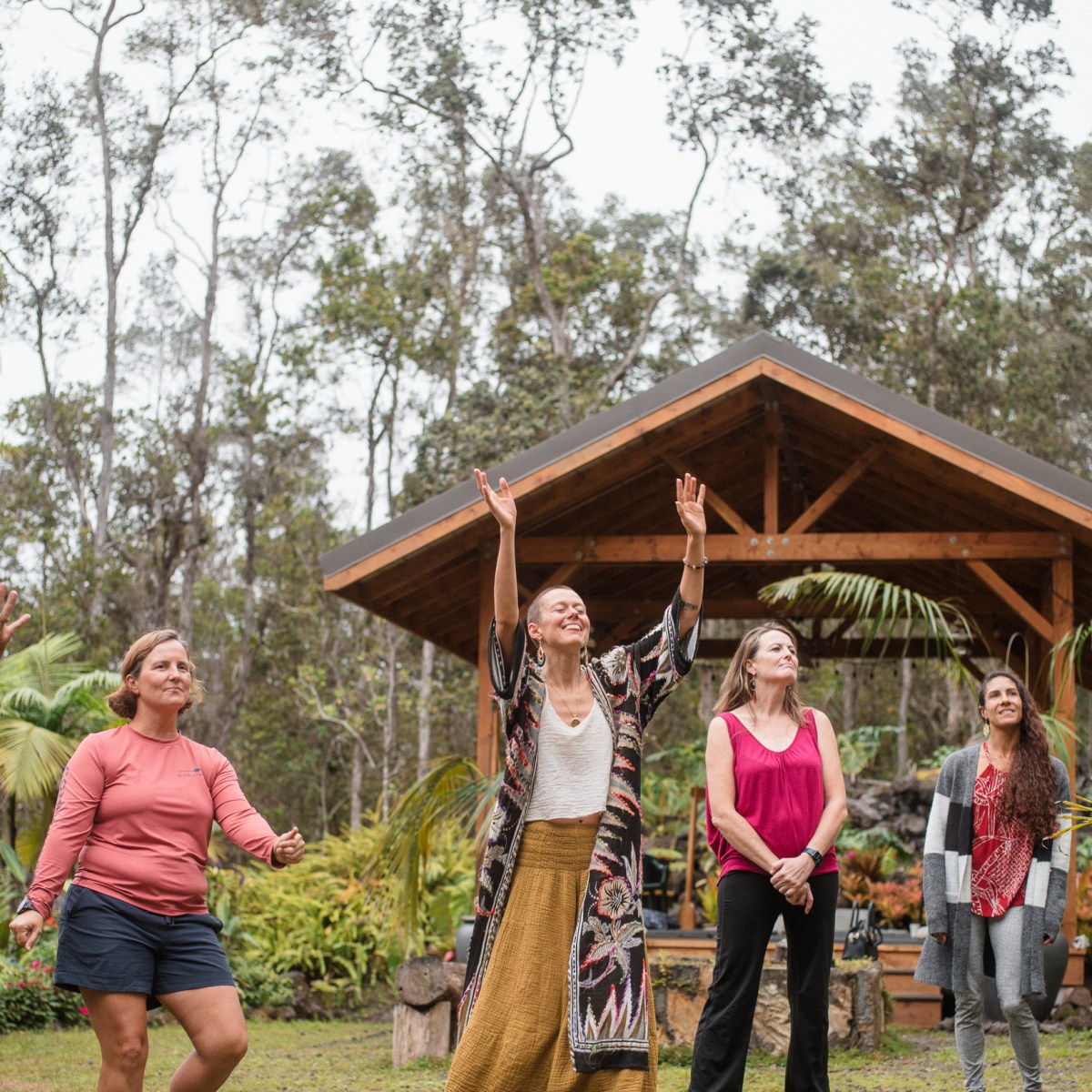 Five people dancing joyfully outdoors near a wooden gazebo.