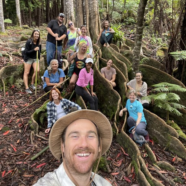 Group of people sitting on tree roots in a forest, smiling at the camera.