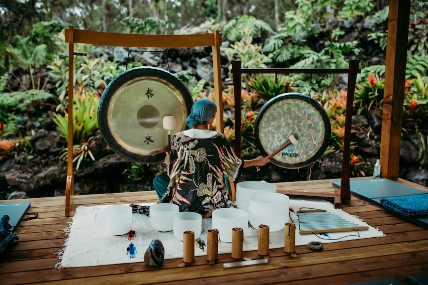 Person playing gongs and bowls outdoors on a wooden platform.