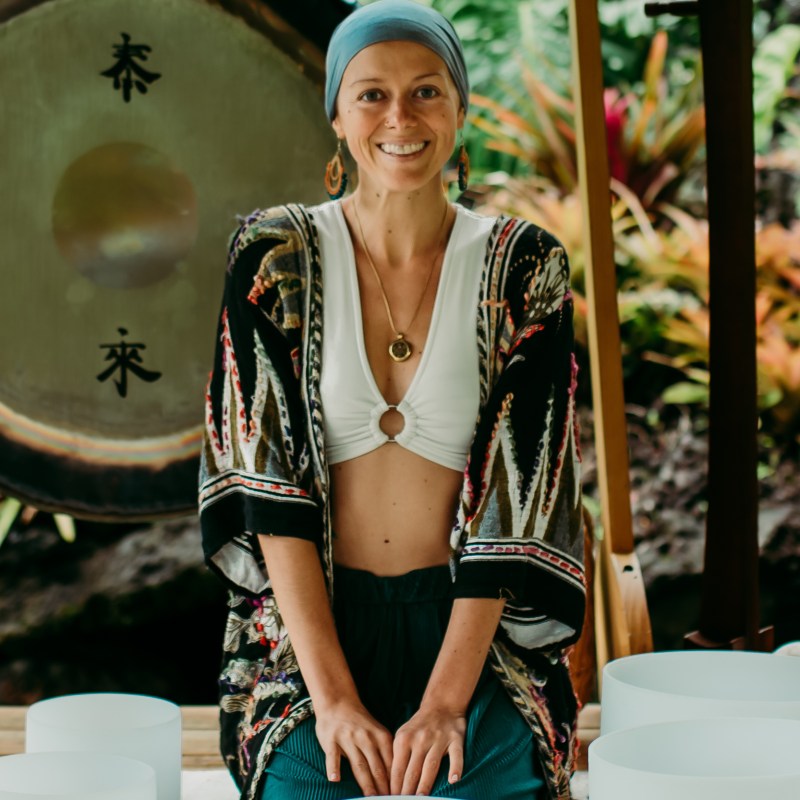 Person in a headscarf with a gong and drums in a lush outdoor setting.
