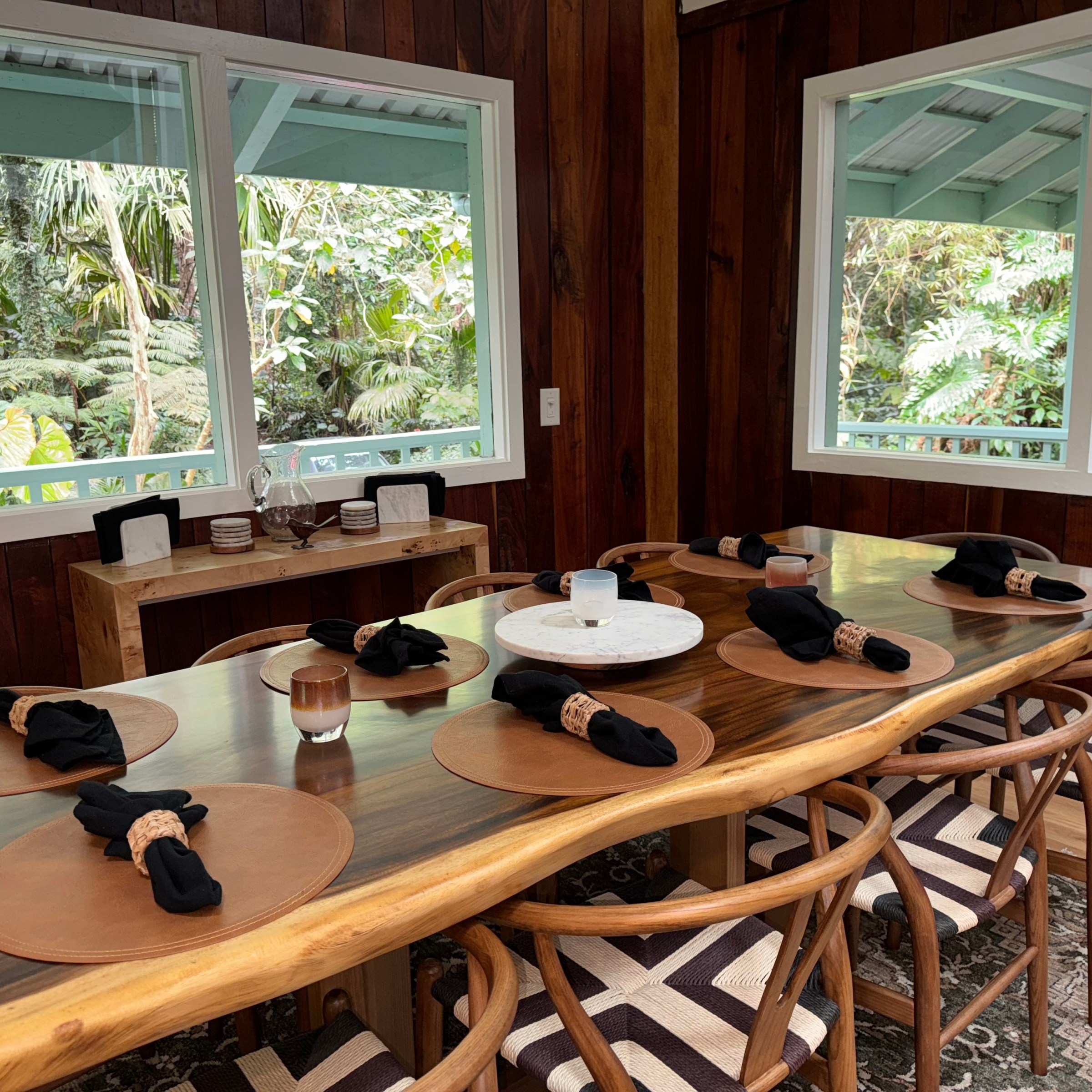 Wooden dining table with eight chairs, set for a meal, near windows with a view of lush greenery.